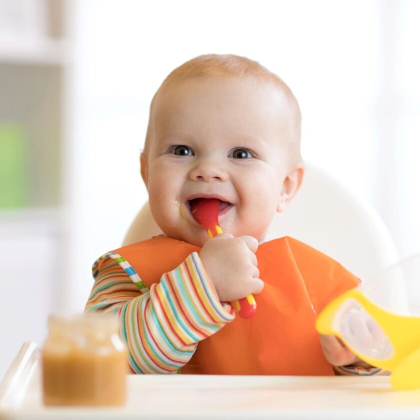 Fr&ouml;hliches Baby isst mit orangefarbenem L&auml;tzchen isst selbstst&auml;ndig Brei aus einer Sch&uuml;ssel.
