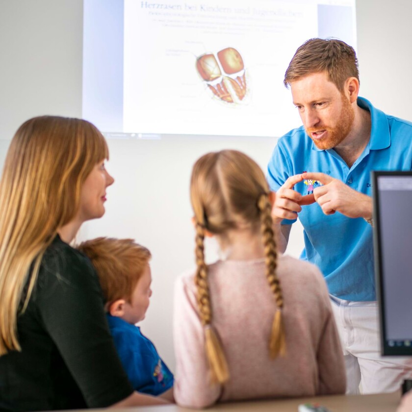 Kinderarzt im blauen Poloshirt erkl&auml;rt Mutter und zwei Kindern gestikulierend Details vor einer Herz-Grafik.