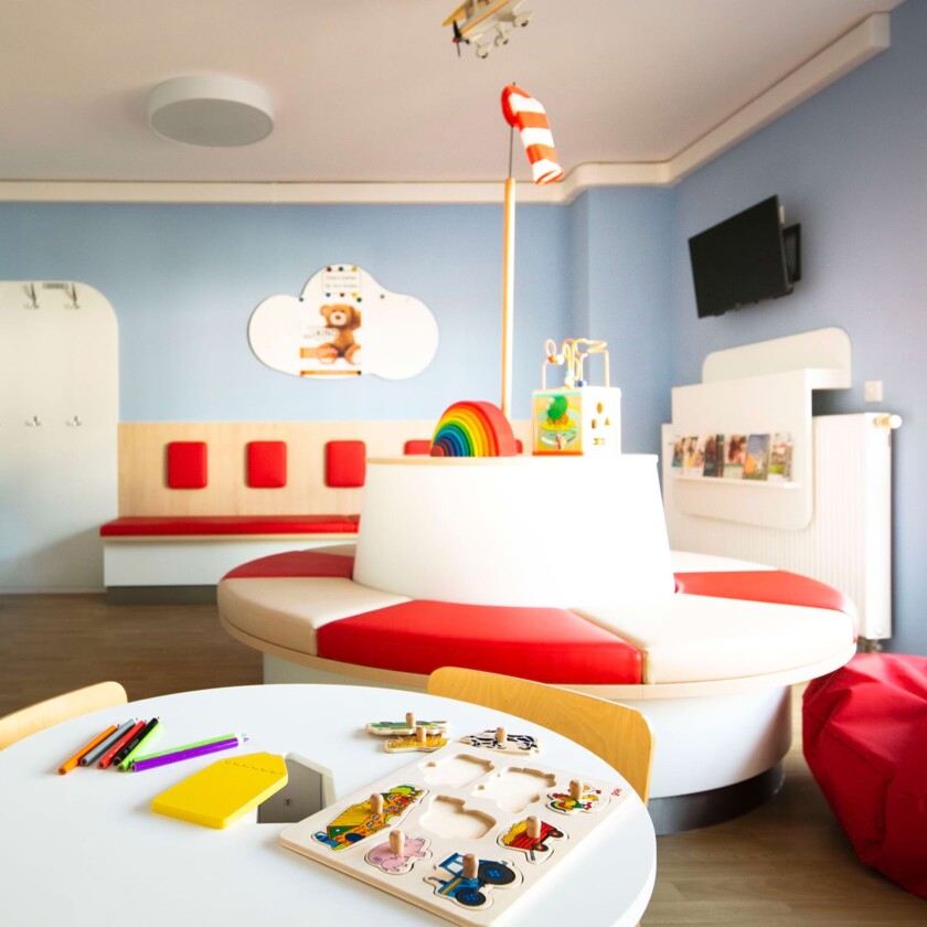 Helles Kinderwartezimmer mit runder rot-wei&szlig;er Sitzinsel, hellblauen W&auml;nden und Spieltisch mit Holzspielzeug.