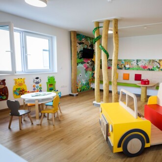 Helles Kinderwartezimmer mit gelbem Holzauto, Spieltisch und farbenfrohen Tiermotiven an der Wand.