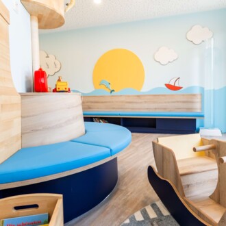 Maritim gestaltetes Kinderwartezimmer mit runder blauer Sitzbank, gro&szlig;em Sonnen-Wandbild und h&ouml;lzernem Spielschiff.