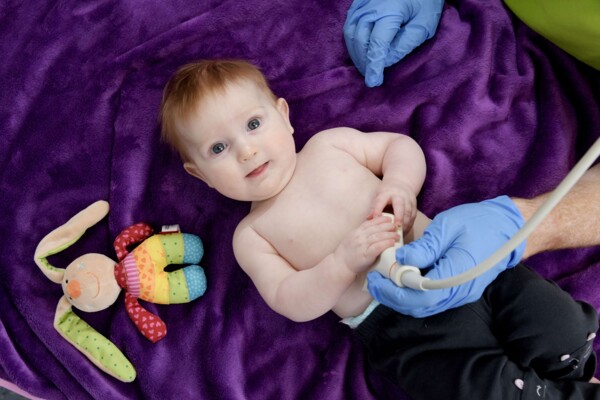 Ruhiges Baby auf violetter Decke wird von Arzt in blauen Handschuhen am Bauch per Ultraschall untersucht.