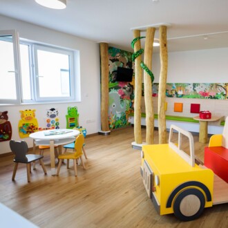 Helles Kinderwartezimmer mit gelbem Holzauto, Spieltisch und farbenfrohen Tiermotiven an der Wand.