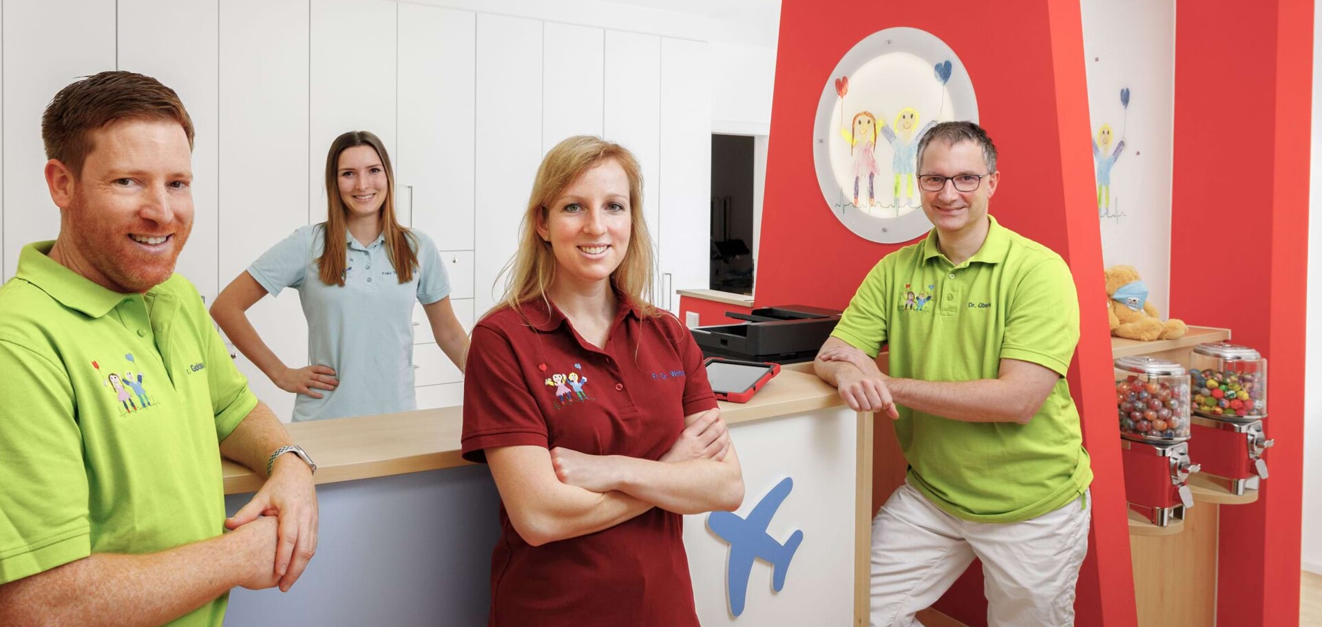 Freundliches Kinderarzt-Team am Empfang: Zwei Mitarbeiterinnen in Hellblau und Rot sowie zwei &Auml;rzte in hellgr&uuml;nen Poloshirts.