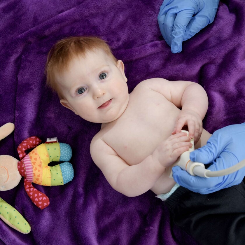 Ruhiges Baby auf violetter Decke wird von Arzt in blauen Handschuhen am Bauch per Ultraschall untersucht.