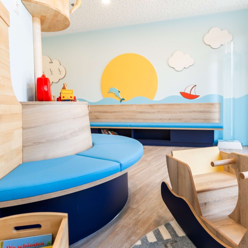 Maritim gestaltetes Kinderwartezimmer mit runder blauer Sitzbank, gro&szlig;em Sonnen-Wandbild und h&ouml;lzernem Spielschiff.