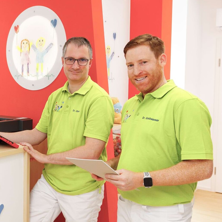 Zwei l&auml;chelnde Kinder&auml;rzte in hellgr&uuml;nen Poloshirts mit Tablet und Dokument in einer bunten Praxis.