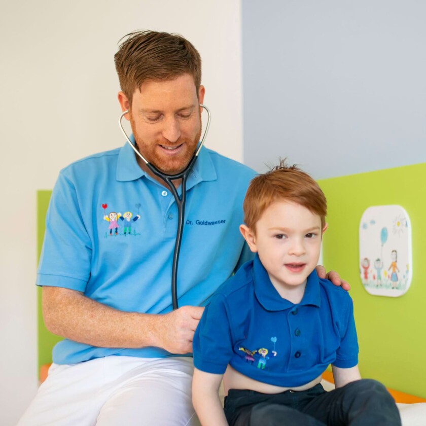 L&auml;chelnder Kinderarzt in hellblauem Poloshirt h&ouml;rt einen kleinen Jungen mit einem Stethoskop ab.