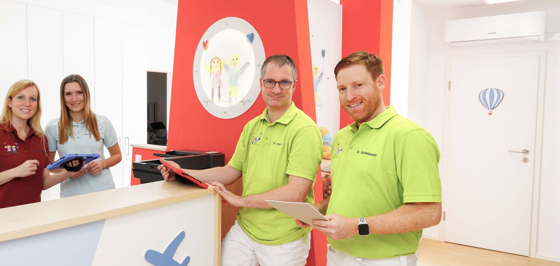 Vierk&ouml;pfiges, l&auml;chelndes Praxisteam in bunten Poloshirts am Empfangstresen einer hellen Kinderarztpraxis.