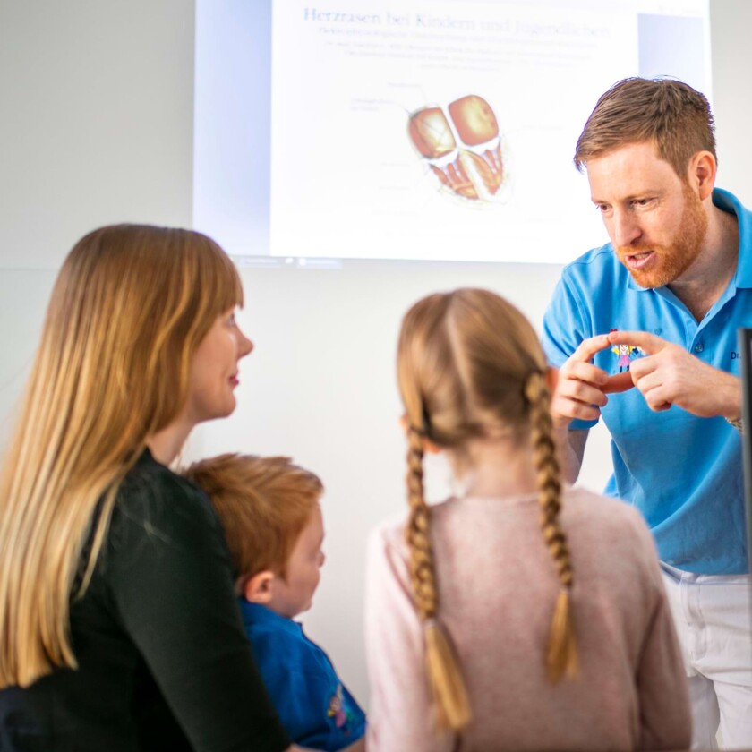 Kinderarzt im blauen Poloshirt erkl&auml;rt Mutter und zwei Kindern gestikulierend Details vor einer Herz-Grafik.