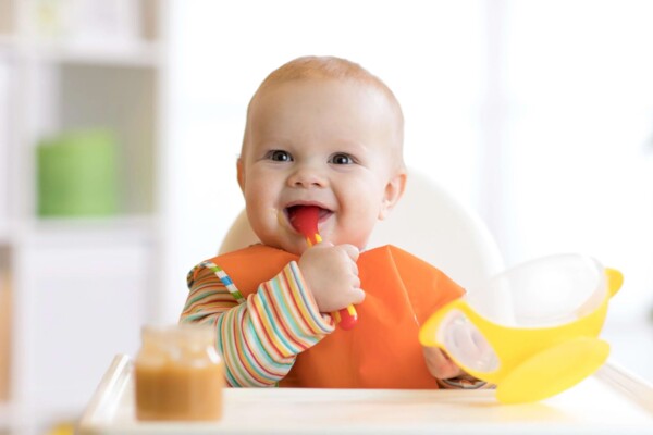 Fr&ouml;hliches Baby isst mit orangefarbenem L&auml;tzchen selbstst&auml;ndig Brei aus einer Sch&uuml;ssel.