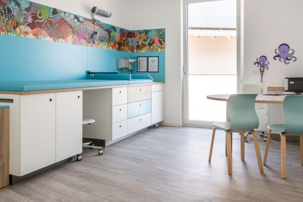 Helles Untersuchungszimmer mit blauer Liege, buntem Unterwasser-Wandbild und rundem Tisch mit hellblauen St&uuml;hlen.