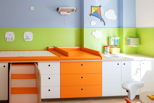 Orange Untersuchungsliege mit ausziehbarer Treppe und Waschbecken vor gr&uuml;n-blauer Wand mit Drachen in einer Praxis.