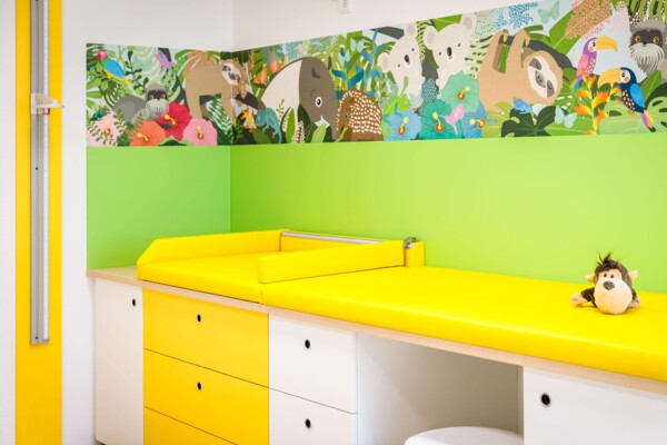 Gelbe Untersuchungsliege mit Pl&uuml;schaffe auf gelb-wei&szlig;en Schr&auml;nken vor gr&uuml;ner Wand mit Dschungelmotiv.