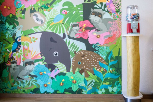 Buntes Dschungel-Wandbild mit Tieren und Bl&uuml;ten neben einem roten Kaugummiautomaten auf einem Holzpodest.