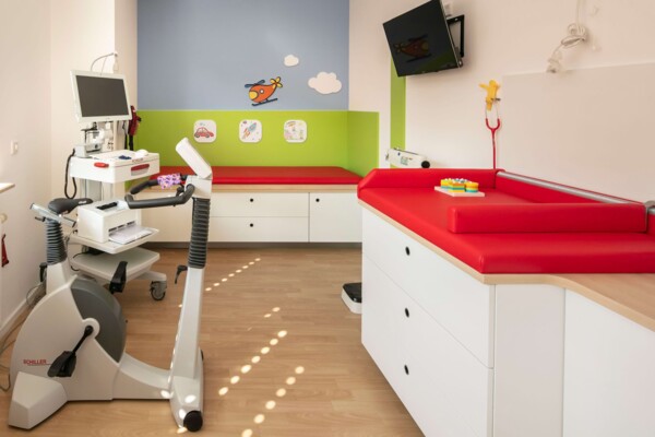 Helles Behandlungszimmer einer Kinderarztpraxis mit Fahrradergometer, roter Liege und bunten Wandmotiven.