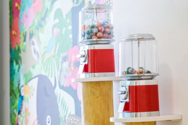 Zwei klassische rote Kaugummiautomaten mit bunten Kugeln auf kleinen Podesten vor einem farbenfrohen Dschungel-Wandbild.