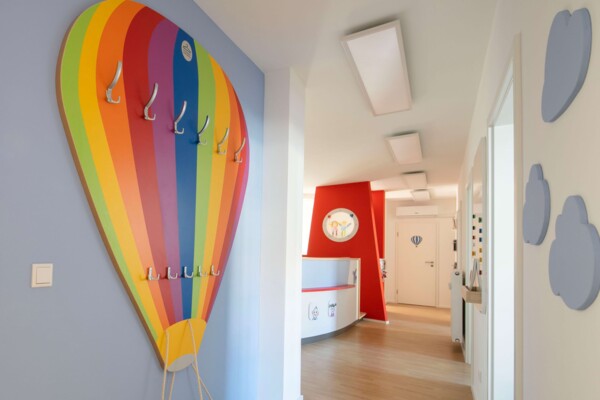 Bunte Garderobe in Form eines Hei&szlig;luftballons an hellblauer Wand im Flur einer hellen Kinderarztpraxis.