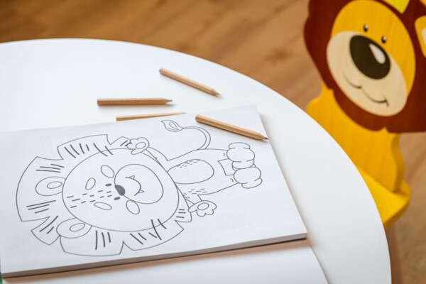 Ausmalbild mit einem L&ouml;wen auf einem wei&szlig;en Kindertisch, daneben Holzstifte und ein gelber Kinderstuhl mit L&ouml;wengesicht.