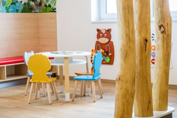 Runder wei&szlig;er Kindertisch mit bunten Tierst&uuml;hlen hinter drei hellen Holzst&auml;mmen in einem freundlichen Wartezimmer.