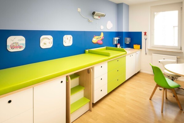 Helles Behandlungszimmer mit gr&uuml;ner Untersuchungsliege, wei&szlig;en Schr&auml;nken und blauer Wand mit fr&ouml;hlichen Kindermotiven.
