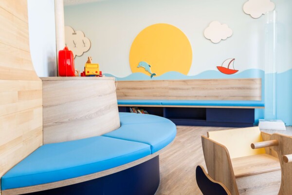 Maritim gestaltetes Kinderwartezimmer mit runder blauer Sitzbank, gro&szlig;em Sonnen-Wandbild und h&ouml;lzernem Spielschiff.