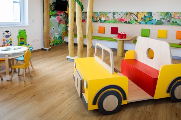 Helles Kinderwartezimmer mit gelb-rotem Holzauto im Vordergrund und buntem Dschungel-Wandbild.