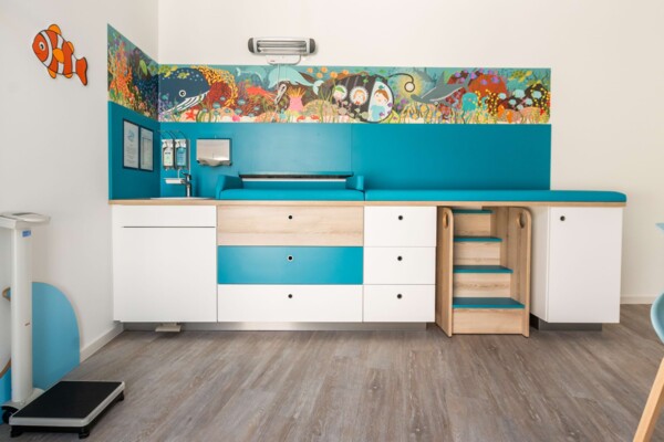 Helles Untersuchungszimmer mit blauer Liege, blau-wei&szlig;en Schr&auml;nken, Holztreppe und buntem Unterwasser-Wandbild.