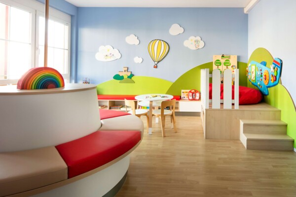 Helles Kinderwartezimmer mit rot-wei&szlig;er Sitzinsel, Holz-Regenbogen und bunter Wandlandschaft mit Hei&szlig;luftballon.