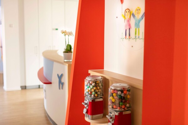 Zwei rote Kaugummiautomaten mit bunten Kugeln vor einer roten Wand im Empfangsbereich einer hellen Kinderarztpraxis.