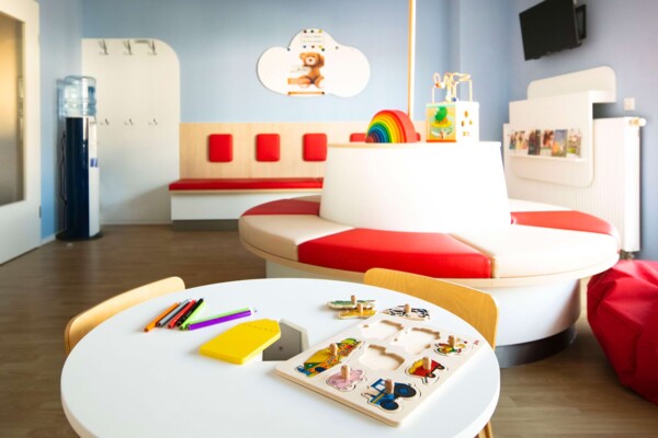 Helles Kinderwartezimmer mit runder rot-wei&szlig;er Sitzinsel, hellblauen W&auml;nden und Spieltisch mit Holzspielzeug.