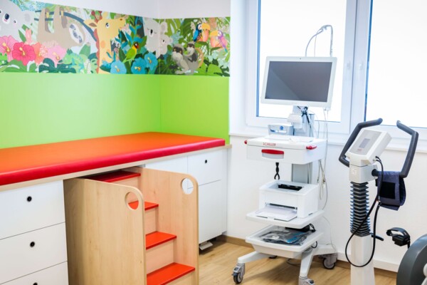 Helles Behandlungszimmer mit roter Liege, Holztreppe, Fahrradergometer und gr&uuml;ner Wand mit buntem Dschungelmotiv.