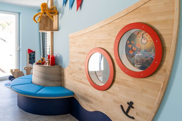 Holzschiff-Spielbereich mit blauer Sitzbank, roten Bullaugen und einem gelben Pl&uuml;sch-Oktopus am Mast.